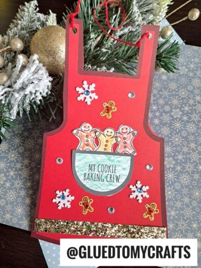 A red paper christmas apron ornament decorated with gingerbread men, snowflakes, and glitter, labeled “MY COOKIE BAKING CREW.” Holiday greenery and gold ornaments are in the background. The handle includes a red string for hanging.