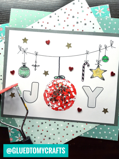 A handmade Christmas card spells “JOY” with a potato masher stamped JOY, the “O” shaped as a red ornament. The letters are decorated with hanging festive items, stars, hearts, and glitter over patterned holiday papers.