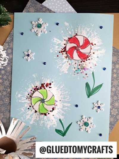 A blue sheet with painted white bursts, decorated with hand-drawn red and green peppermint candy flowers, snowflake stickers, glitter, and green painted leaves. The username @GLUEDTOMYCRAFTS is at the bottom.