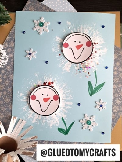 A handmade winter card featuring two smiling snowman faces, each crafted with a charming toilet roll stamped snowman design, painted dandelion-like backgrounds, snowflake stickers, sequins, green leaves, and the text @GLUEDTOMYCRAFTS at the bottom.