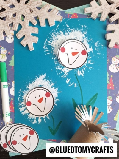 A blue paper features toilet roll stamped snowman faces made from white paint circles and paper cutouts. Snowflake decorations, a green marker, and a cardboard tube with cut slits are nearby. @GLUEDTOMYCRAFTS logo at the bottom.
