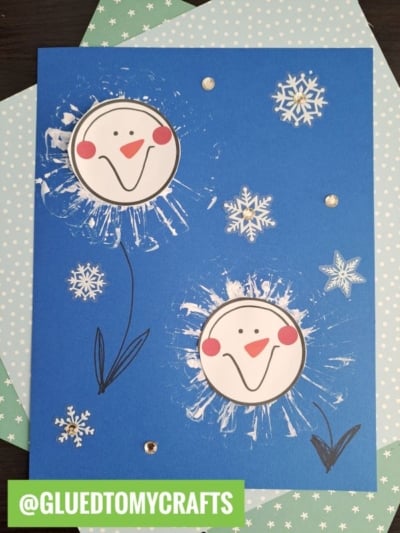A blue card features two smiling toilet roll stamped snowman faces with hand-drawn eyes and orange noses, surrounded by white paint splatters, paper snowflakes, and sequins. The handle @GLUEDTOMYCRAFTS is displayed at the bottom left.
