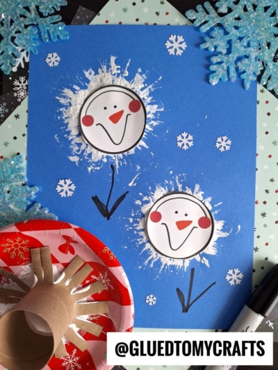 Blue paper features two toilet roll stamped snowman faces, made with white paint and finished with drawn arms and smiles. Snowflake decorations and crafting materials are scattered around. @GLUEDTOMYCRAFTS is at the bottom.