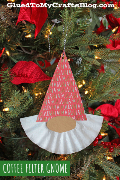 A handmade coffee filter Christmas gnome ornament, featuring a patterned red paper hat, light brown nose, and white coffee filter beard, hangs festively on a Christmas tree adorned with red poinsettias and glowing yellow lights.