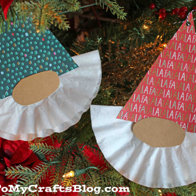 Two handmade coffee filter Christmas gnome ornaments, featuring patterned paper hats, oval noses, and white ruffled beards, hang on a decorated Christmas tree adorned with red berries and twinkling lights.