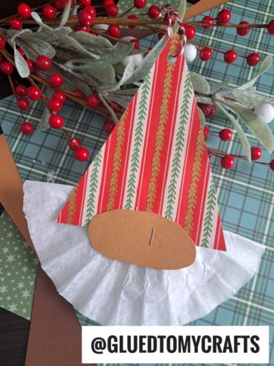 A handmade coffee filter Christmas gnome ornament featuring a paper coffee filter beard, patterned paper hat, and brown paper nose. The handle @GLUEDTOMYCRAFTS appears at the bottom, with festive decor in the background.