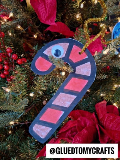 A paper candy cane reindeer ornament with red and white stripes and a googly eye hangs on a Christmas tree decorated with red berries and poinsettias. The handle @GLUEDTOMYCRAFTS is displayed at the bottom.