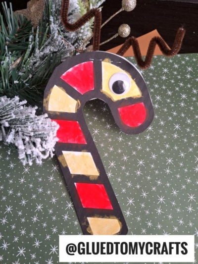 A candy cane reindeer paper craft, decorated with red and yellow sections, a large googly eye, and brown pipe cleaner antlers, is displayed on a green star-patterned background next to frosted faux greenery.