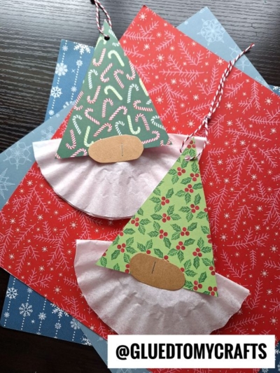 Two handmade coffee filter Christmas gnome ornaments crafted from patterned paper, coffee filters, and string rest on red and blue snowflake-patterned paper. The image features the text @GLUEDTOMYCRAFTS.