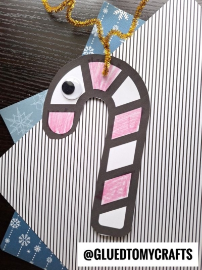 A handmade candy cane reindeer ornament with a black outline, pink and white sections, a googly eye, and a gold pipe cleaner loop on top, displayed on striped and patterned paper. The text @GLUEDTOMYCRAFTS is at the bottom.