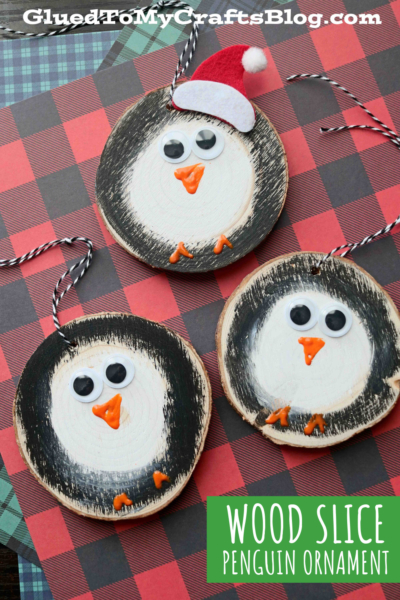 Three handmade wood slice penguin ornaments feature painted faces, googly eyes, and orange beaks. One penguin sports a red Santa hat. All are displayed on a charming plaid background.