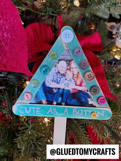 A family photo is framed in a green triangle made of craft sticks decorated with colorful buttons, hanging on a Christmas tree as a Cute As A Button Ornament. The words CUTE AS A BUTTON are written at the bottom.