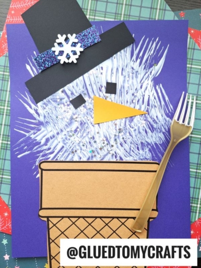 A fork painted snowman craft made from paper, featuring a textured white face, black hat, orange triangle nose, brown ice cream cone body, and plastic fork on a purple background. @GLUETOMYCRAFTS is written at the bottom.