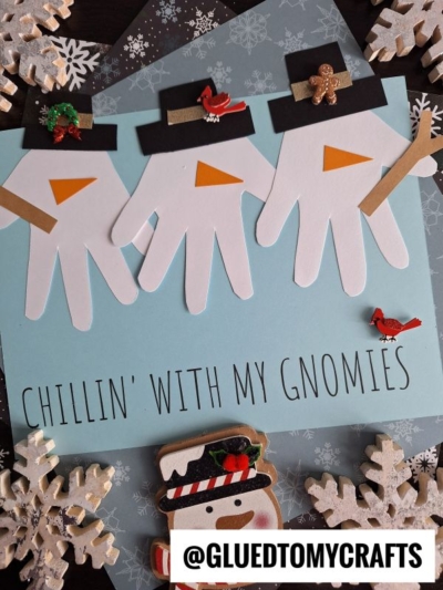 Handprint cutouts decorated as snowman gnomes with hats and noses on blue paper, surrounded by snowflakes, holiday decorations, and a snowman cookie. Text reads “Chillin’ with my gnomies” and “@GLUEDTOMYCRAFTS.”.