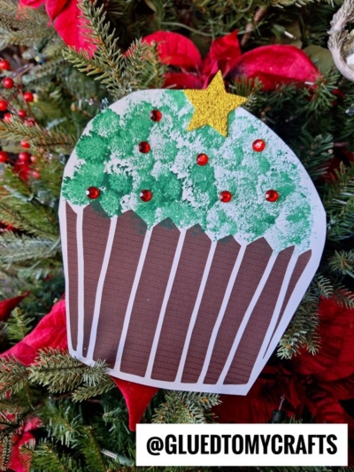 A handmade Christmas tree cupcake ornament with green frosting, red dots, and a yellow star topper hangs on a festive tree. The brown and white striped liner stands out among red poinsettias and pine needles. @GLUEDTOMYCRAFTS is at the bottom.