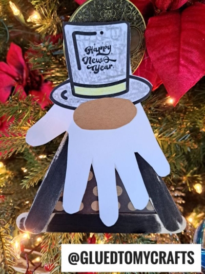 A Popsicle Stick New Year's Eve Gnome ornament shaped like a person with a top hat that says “Happy New Year,” made from paper cutouts, hangs on a Christmas tree. The handle @GLUEDTOMYCRAFTS is shown at the bottom.