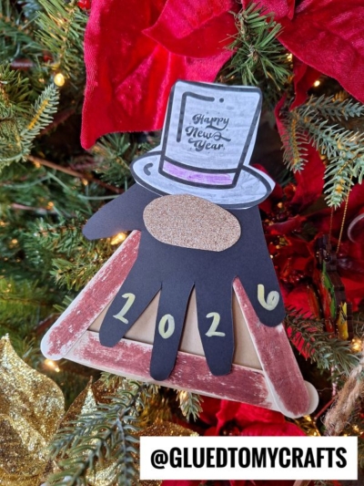 A handmade Popsicle Stick New Year’s Eve Gnome ornament hangs on a Christmas tree. It features a black paper hand, a glittery top hat with Happy New Year, 2024 numbers, and an @GLUEDTOMYCRAFTS sign at the bottom.