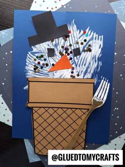 A fork painted snowman craft made with white paint, a brown paper pot, black hat, orange nose, and colorful dots on blue paper. “@GLUEDTOMYCRAFTS” is displayed at the bottom.