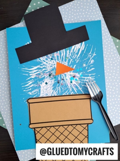 A fork painted snowman craft made from colored paper features a black hat, orange triangle nose, and is displayed on a blue background with snowflake designs. The image includes the handle @GLUETOMYCRAFTS.