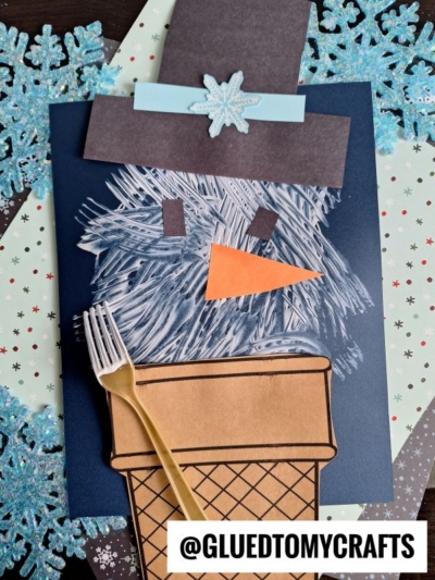 A fork painted snowman made from blue, black, and brown paper features a large orange carrot nose and top hat, decorated with a blue snowflake. A plastic fork rests nearby on a wintry background. @GLUEDTOMYCRAFTS is at the bottom.