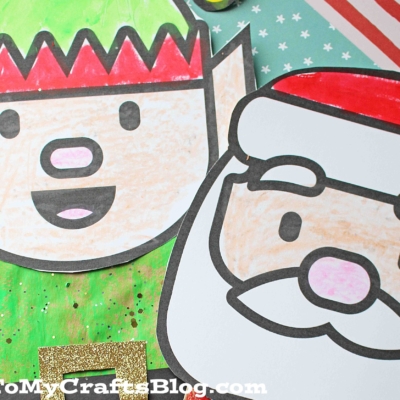 Two paper bag puppets—a paper bag elf and a paper bag Santa—decorated with colored paper and glitter belts, lie on a striped background.