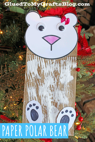 A paper bag polar bear, complete with googly eyes, a pink bow, pink nose, and white painted fur, is displayed on a Christmas tree. The text below reads “Paper Polar Bear.”.