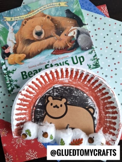 A children’s book, Bear Stays Up, sits on holiday-themed paper. Below is a festive paper plate craft with a bear face, red paint, cotton balls, small acorn stickers, and a cozy paper plate bear cave for added fun. @GLUEDTOMYCRAFTS is at the bottom.