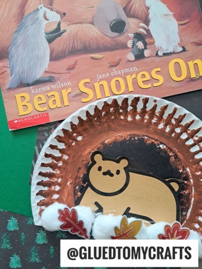 A children’s book titled “Bear Snores On” is next to a paper plate bear cave craft decorated with cotton balls and paper leaves. The username @GLUEDTOMYCRAFTS appears at the bottom.