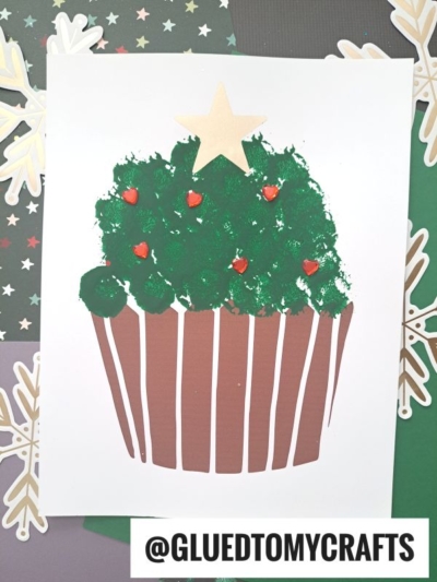 Children’s Christmas craft: A Christmas tree cupcake made by dabbing green paint into a cupcake shape with a brown base and a gold star on top, decorated with red dots resembling holly berries. The background features snowflake and star-themed paper.
