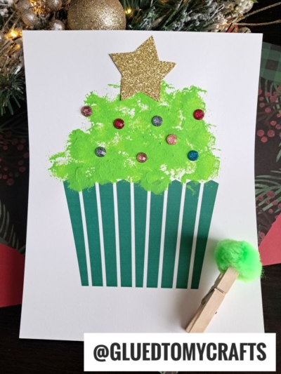 A Christmas tree cupcake craft featuring green pom-pom frosting, colorful round decorations, and a glittery gold star on top. A green pom-pom attached to a clothespin sits beside the festive treat.