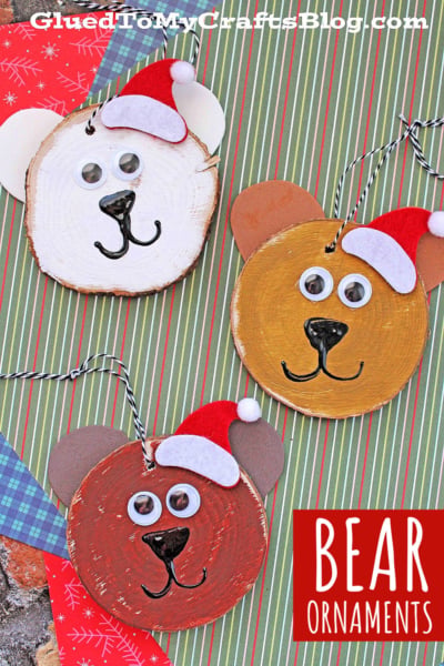 Three bear-shaped Christmas ornaments, each a charming wood slice bear ornament with googly eyes, a drawn-on nose and mouth, and a tiny red Santa hat. The ornaments are displayed on striped and patterned paper.