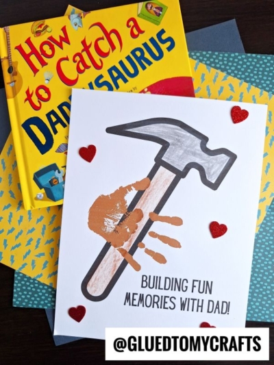 A children’s craft features a handprint hammer on paper, surrounded by red glitter hearts. Above it is the book How to Catch a Daddysaurus. Text reads: BUILDING FUN MEMORIES WITH DAD!.