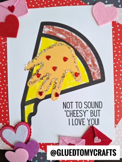 A handmade Valentines card features a playful handprint pizza slice as the centerpiece, decorated with red heart stickers and glitter. The red, heart-filled background frames the text: Not to sound cheesy but I love you!.