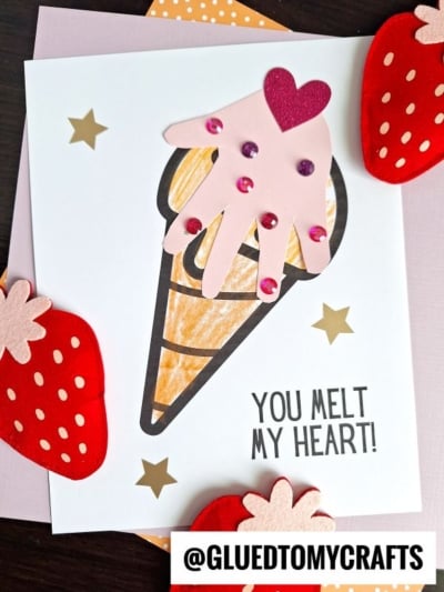 A handmade handprint valentine card features an ice cream cone with a scoop made from a pink paper handprint, decorated with sequins and a paper heart. The card reads You melt my heart! and is surrounded by red strawberry cutouts.