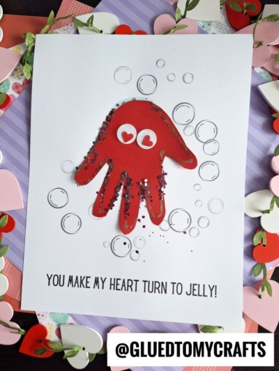 A handmade card features a red handprint jellyfish with googly eyes and glitter. Drawn bubbles float around it, and below, text reads, You make my heart turn to jelly! The scene is surrounded by charming paper hearts.
