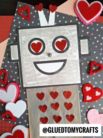 A handmade paper bag robot with a gray, square head, heart-shaped eyes, and a smiling mouth, surrounded by red and white heart decorations on a colorful, heart-patterned background.