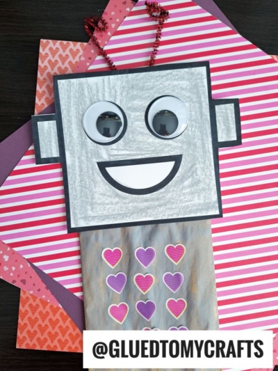 A handmade paper bag robot craft made from a gray bag with heart stickers, large googly eyes, a smiling mouth, and pipe cleaner antennae, displayed on colorful striped and heart-patterned paper.