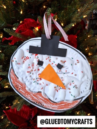 A puffy paint snowman craft made from paper and white textured material hangs on a Christmas tree, decorated with red and green sprinkles like a donut, a paper carrot nose, black hat, black eyes, and a pink ribbon for hanging.
