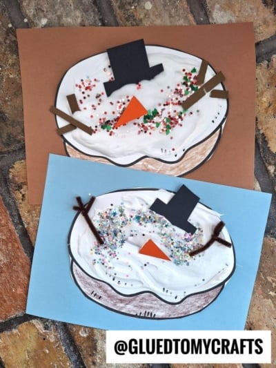 Two puffy paint snowman face crafts made from paper, white paint, and colorful glitter. Each has a black hat, orange carrot nose, brown stick arms, and sits on blue and brown backgrounds—like a frosty snowman donut treat! @GLUEDTOMYCRAFTS.