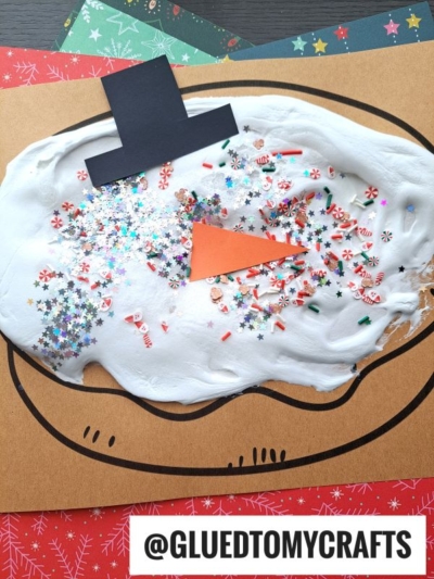 A puffy paint snowman face craft made with white glue, glitter, and confetti on brown paper. It features a black paper hat, orange carrot nose, and is surrounded by colorful festive paper. @GLUEDTOMYCRAFTS is written at the bottom.