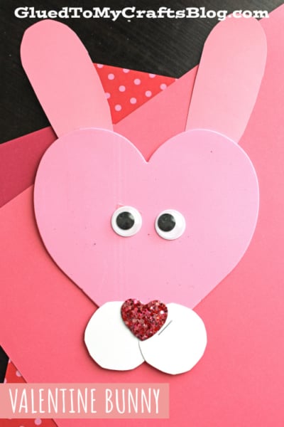 A pink heart-shaped paper bunny craft with googly eyes, two pink ears, white cheeks, and a glittery red heart nose sits on red and pink paper. Perfect for heart shaped crafts or valentine art project ideas. Text reads “VALENTINE BUNNY.”.