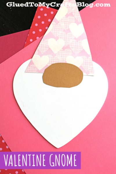 A Valentine gnome craft made with a large white craft foam heart for the body, a tan oval nose, and a pink hat decorated with white hearts, placed on pink and red paper—perfect for heart shaped crafts or valentine art project ideas.