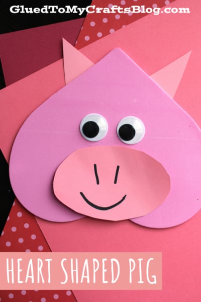 A heart shaped pig made from pink and red paper with googly eyes, a round snout, and a smiling mouth—perfect for heart shaped crafts or valentine art project ideas. Displayed on a red and pink background. Text reads HEART SHAPED PIG.