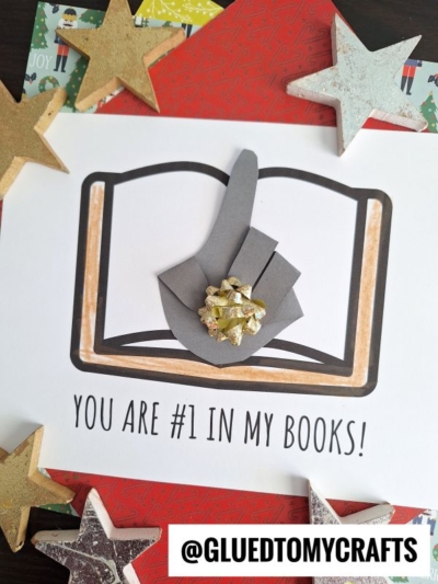 A handmade card with a paper hand showing a number one gesture over an open handprint book illustration. Text below reads, You are #1 in my books! Gold and white stars decorate the card.