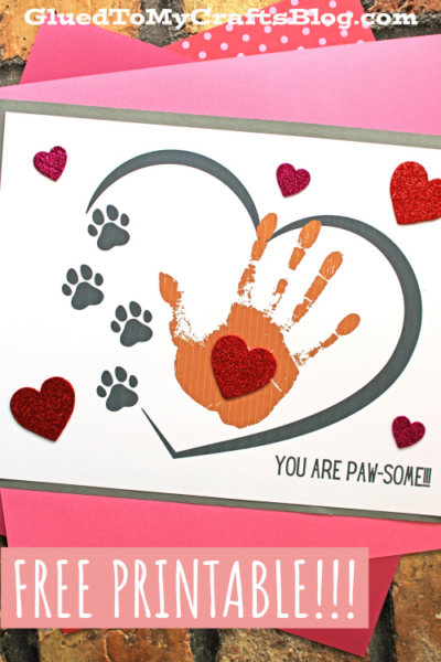 A handmade card featuring a brown handprint, gray paw prints, red glitter hearts, and a large outlined heart. The text “you are paw-some” and “FREE PRINTABLE!!!” pop on a pink background.