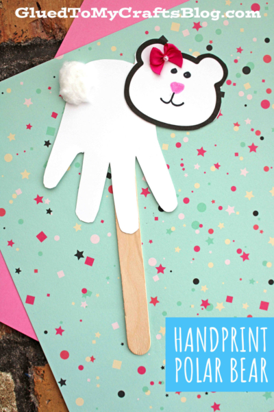 A handmade handprint polar bear craft features a white paper handprint as the body, a drawn bear face, pink bow, white pom-pom ear, and popsicle stick handle, all set against colorful patterned paper.