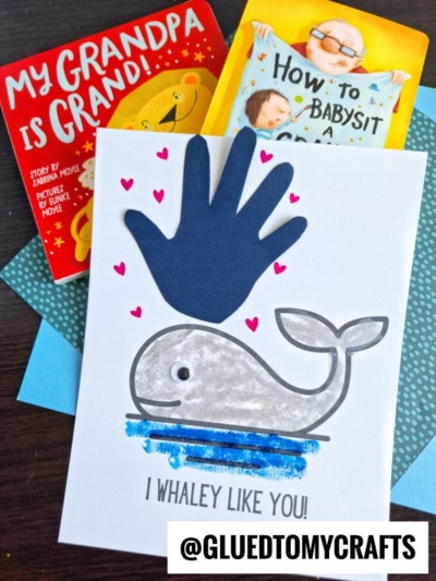 Greeting card featuring a handprint whale with gray body, blue lines, googly eye, and pink hearts. Above it is a dark blue paper handprint. In the background are books: My Grandpa is Grand! and How to Babysit a Grandpa.