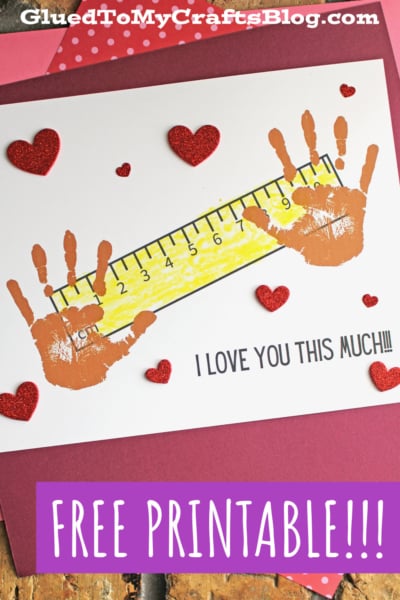 A handmade Valentine’s card says “love you this much,” featuring two brown handprints, a yellow ruler between them, and red glitter hearts. The text reads, “I LOVE YOU THIS MUCH!!!” plus a “FREE PRINTABLE!”.