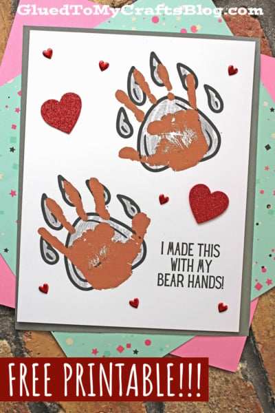 A handmade card features two brown handprint bear hands outlined in black to resemble bear paws, red glitter heart stickers, and the text I MADE THIS WITH MY BEAR HANDS! on white paper. The card sits on colored paper.