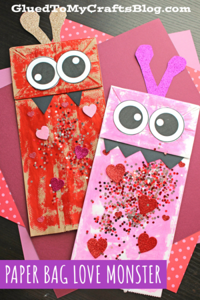 Two paper bag love monsters—one red and one pink—feature googly eyes, heart shapes, glitter, and jagged black mouths, displayed atop layered pink and red paper backgrounds.